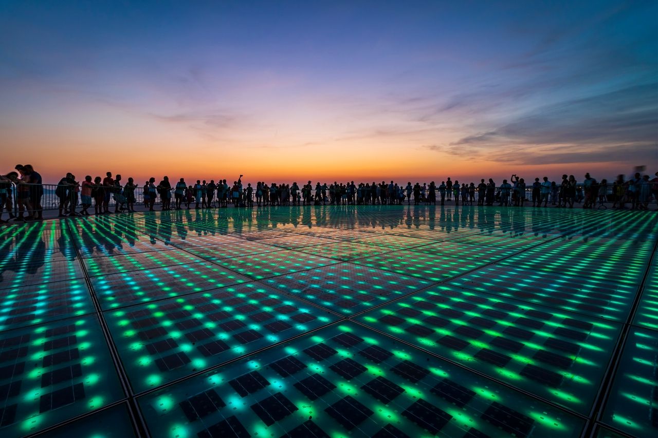 Der "Gruß an die Sonne" zeigt, eine große kreisförmige Installation mit farbigen LED-Lichtern im Boden. Viele Menschen stehen um die Installation herum und genießen die Lichtershow.