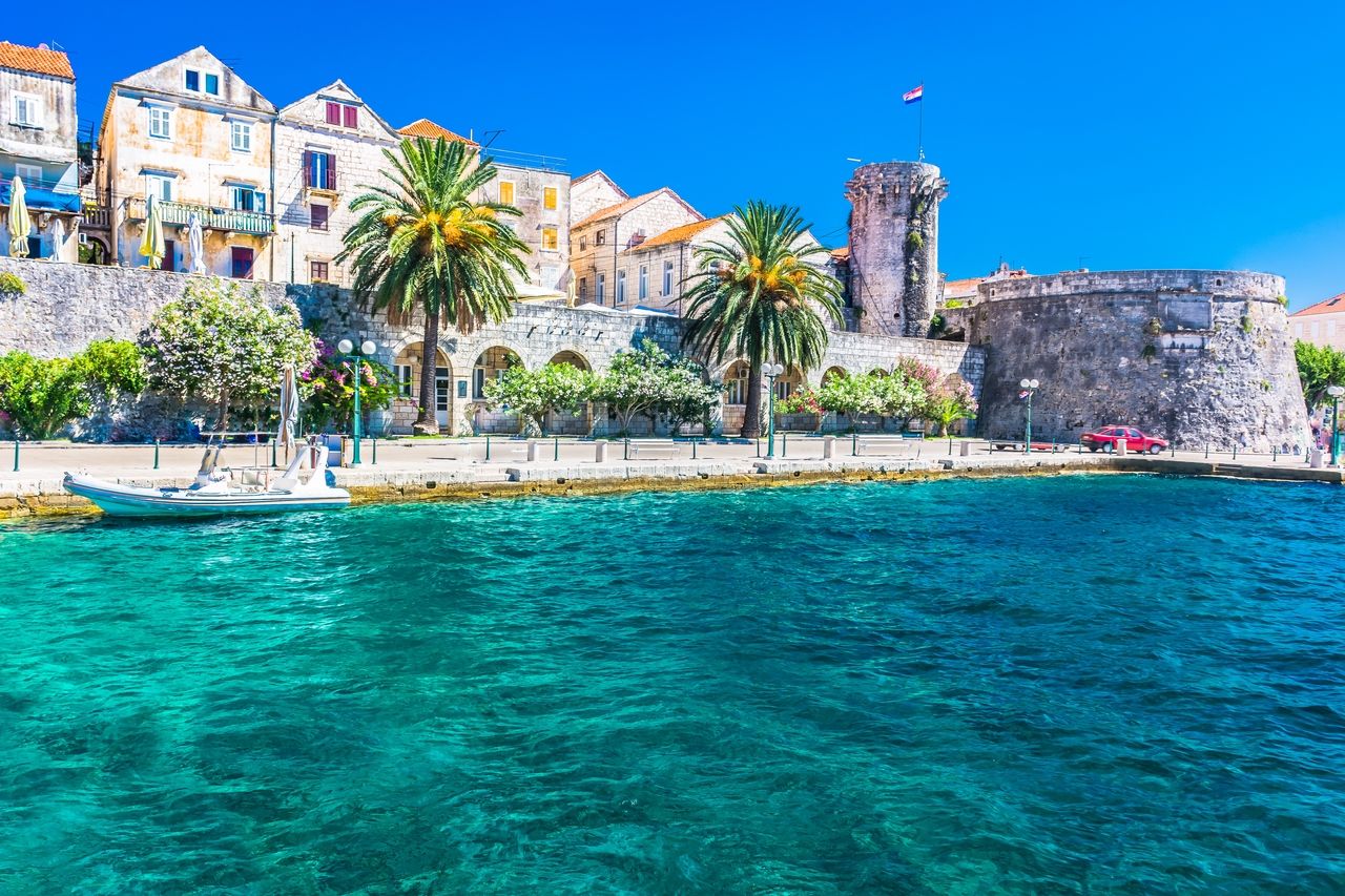 Küste von Korčula mit historischen Gebäuden und einer beeindruckenden Steinmauer im Hintergrund. Die Uferpromenade ist gesäumt von Palmen und blühenden Pflanzen. Im Vordergrund schimmert das kristallklare blaue Wasser der Adria.