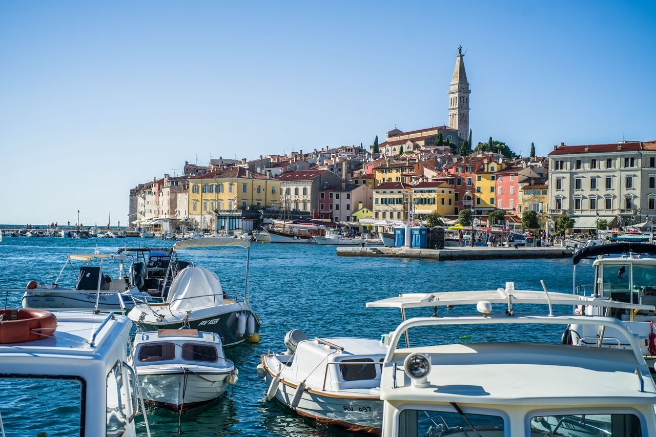 Der Hafen von Rijeka, mit vielen Booten im Vordergrund, die im klaren blauen Wasser schwimmen. Im Hintergrund ist die historische Altstadt von Rijeka zu sehen, mit bunten Häusern und der markanten Kirche, die auf einem Hügel thront.