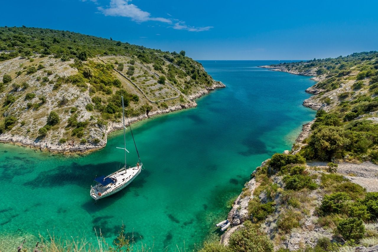 Inlet with turquoise water and a sailboat, surrounded by green hills.