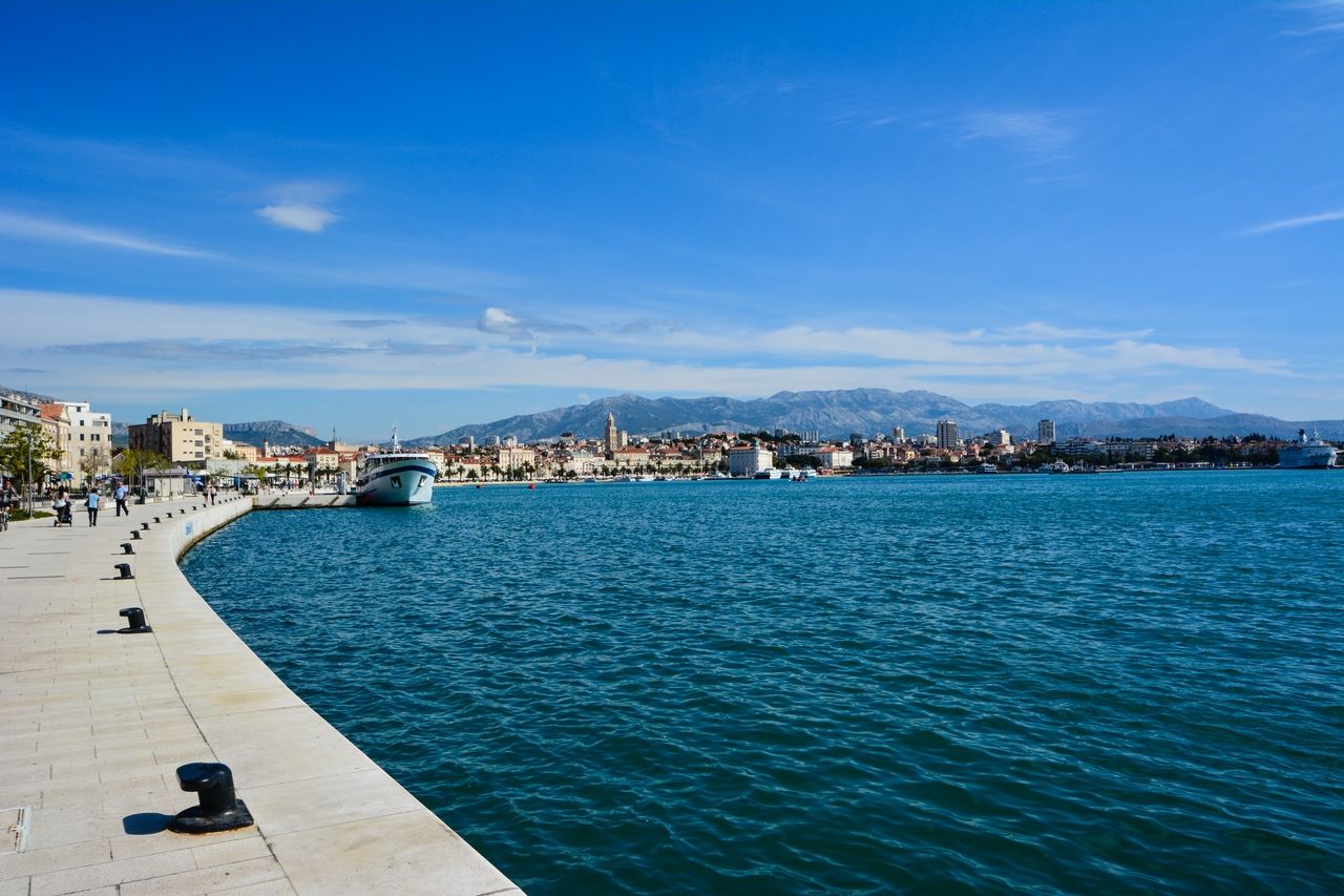 Ein weiter Blick auf die Uferpromenade von Split mit einem großen Schiff, das im Hafen angelegt hat. Die Promenade ist mit Spaziergängern belebt und führt entlang des tiefblauen Wassers. Im Hintergrund sind die historische Stadt Split und die umliegenden Berge unter einem klaren blauen Himmel zu sehen.