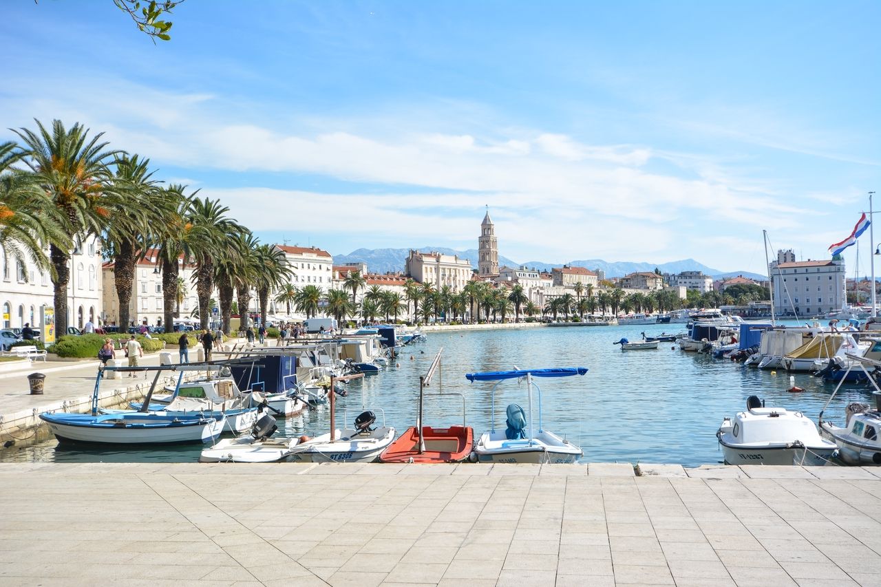 Ein malerischer Blick auf die Uferpromenade von Split, gesäumt von hohen Palmen und historischen Gebäuden. Im Vordergrund sind mehrere Boote im ruhigen Wasser des Hafens zu sehen. Im Hintergrund ragt der Glockenturm des Diokletianpalastes empor und die umliegenden Berge bieten eine beeindruckende Kulisse.