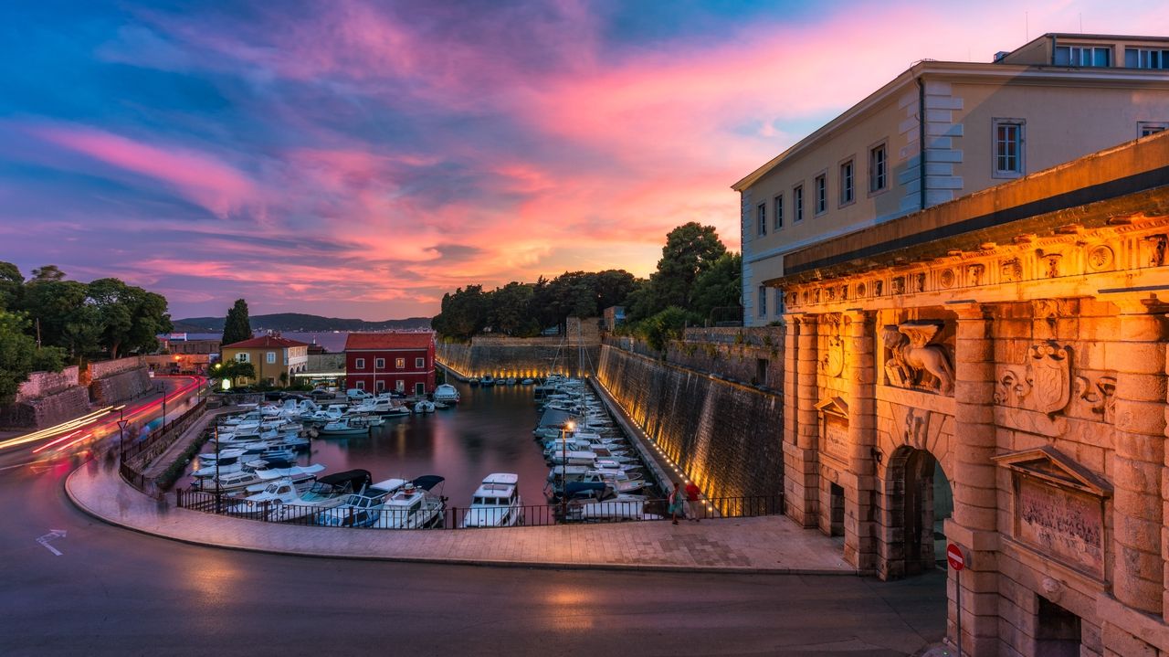 Zadar bei Sonnenuntergang mit einem kleinen Hafen voller Boote, umgeben von historischen Mauern und Gebäuden. Die warmen Lichter der Stadt reflektieren auf dem Wasser, während der Himmel in rosa und violetten Farbtönen leuchtet. Ein historisches Tor ist im Vordergrund zu sehen, beleuchtet von orangefarbenem Licht.