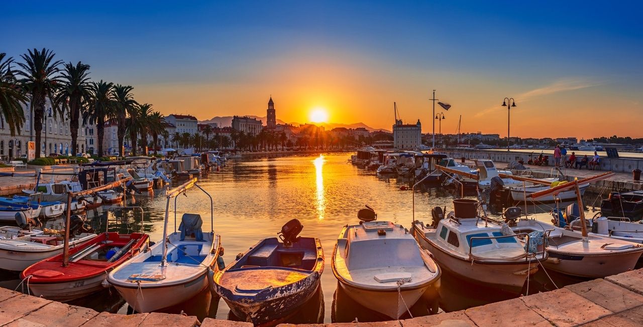 Solnedgangen over den maleriske havnepromenade i Split i Kroatien, omgivet af palmer og adskillige både, der ligger fredeligt i vandet. I baggrunden ses historiske bygninger og en kirke, der fremhæver byens kulturelle skønhed.