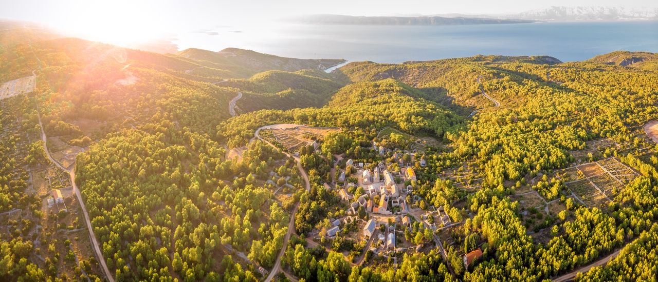 Luchtfoto van het stenen dorp Humac op Hvar bij zonsondergang, omringd door groene heuvels en oude veldmuren, met op de achtergrond de zee en het vasteland.