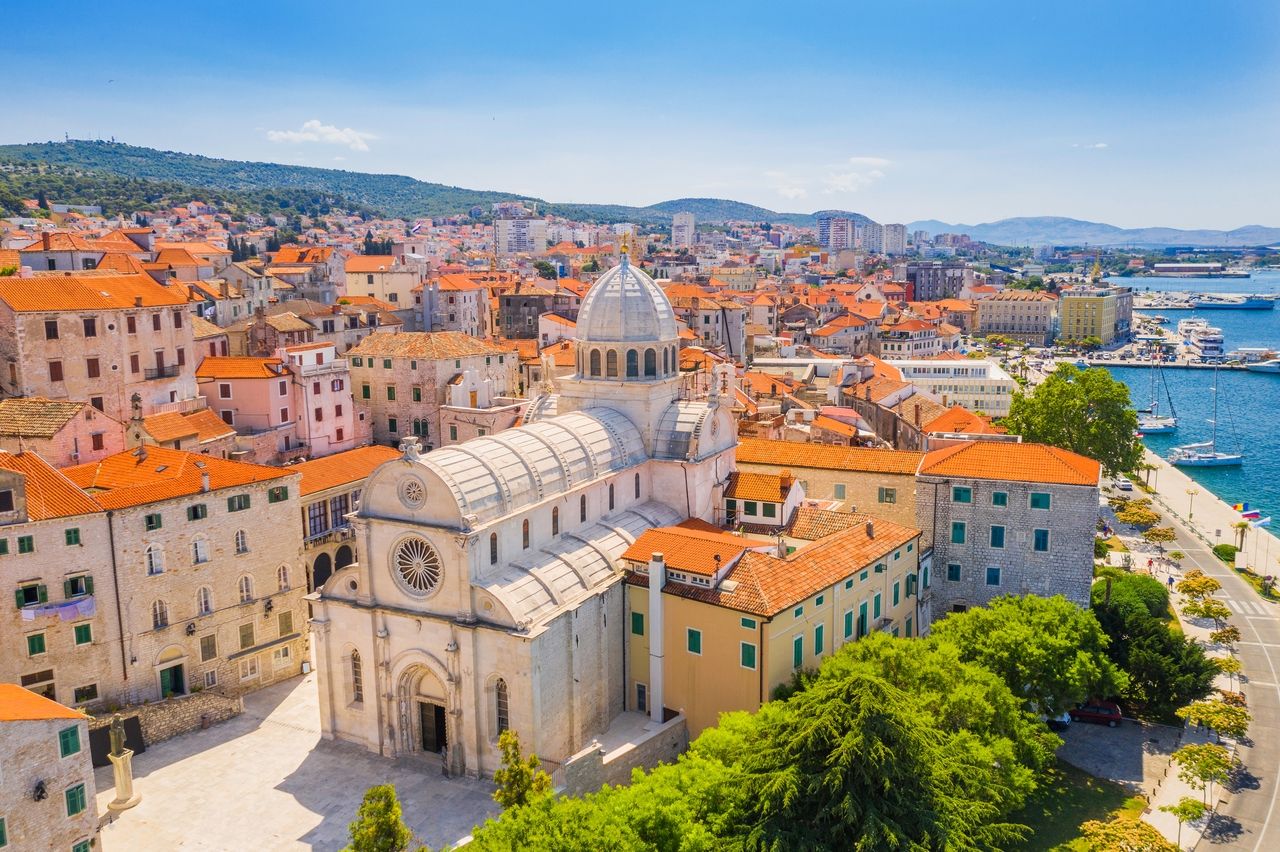 Ön planda belirgin katedrali ile Šibenik'in havadan görünümü. Şehir, geleneksel kırmızı çatılar ve tarihi taş binalarla dikkat çekiyor. Arka planda modern binalar ve yelkenli teknelerle dolu pitoresk liman görülüyor.