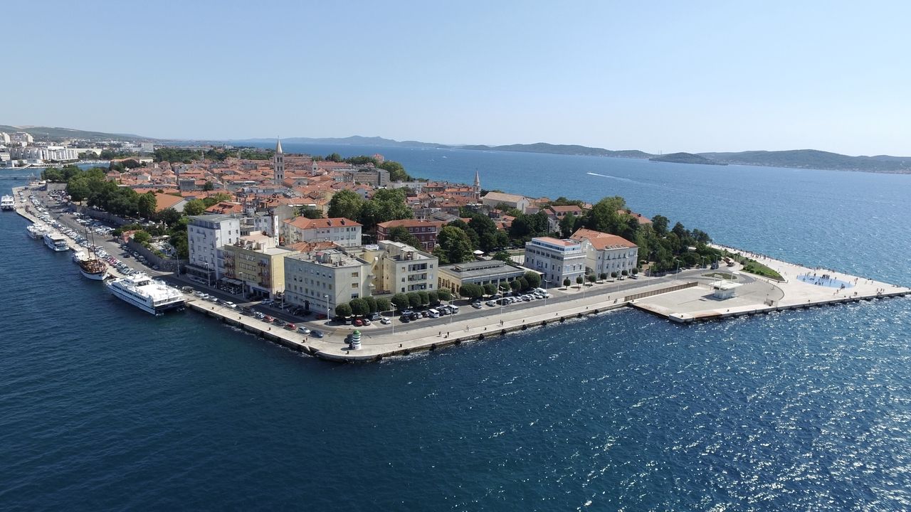 Luftaufnahme der Küstenstadt Zadar, mit Such auf die Altstadt mit ihren historischen Gebäuden und roten Dächern zeigt. Entlang der Uferpromenade sind zahlreiche Boote und Schiffe zu sehen. Im Vordergrund befindet sich der Platz mit der berühmten Meeresorgel und dem Gruß an die Sonne.
