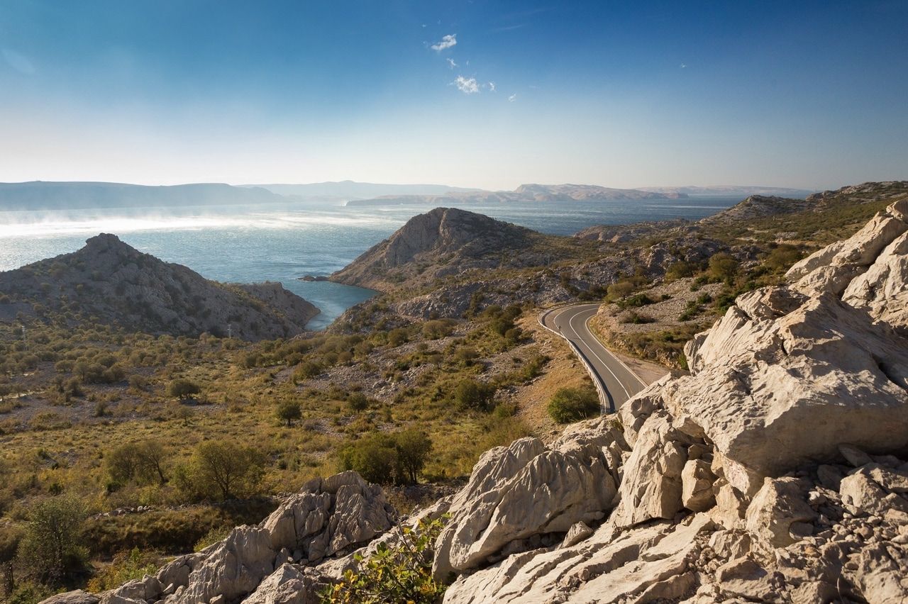 Panorámakép egy kanyargós útról, amely a horvát tengerpart mentén halad, és a sziklás tájon kanyarog. A háttérben az Adriai-tenger csillog a tiszta, kék ég alatt.