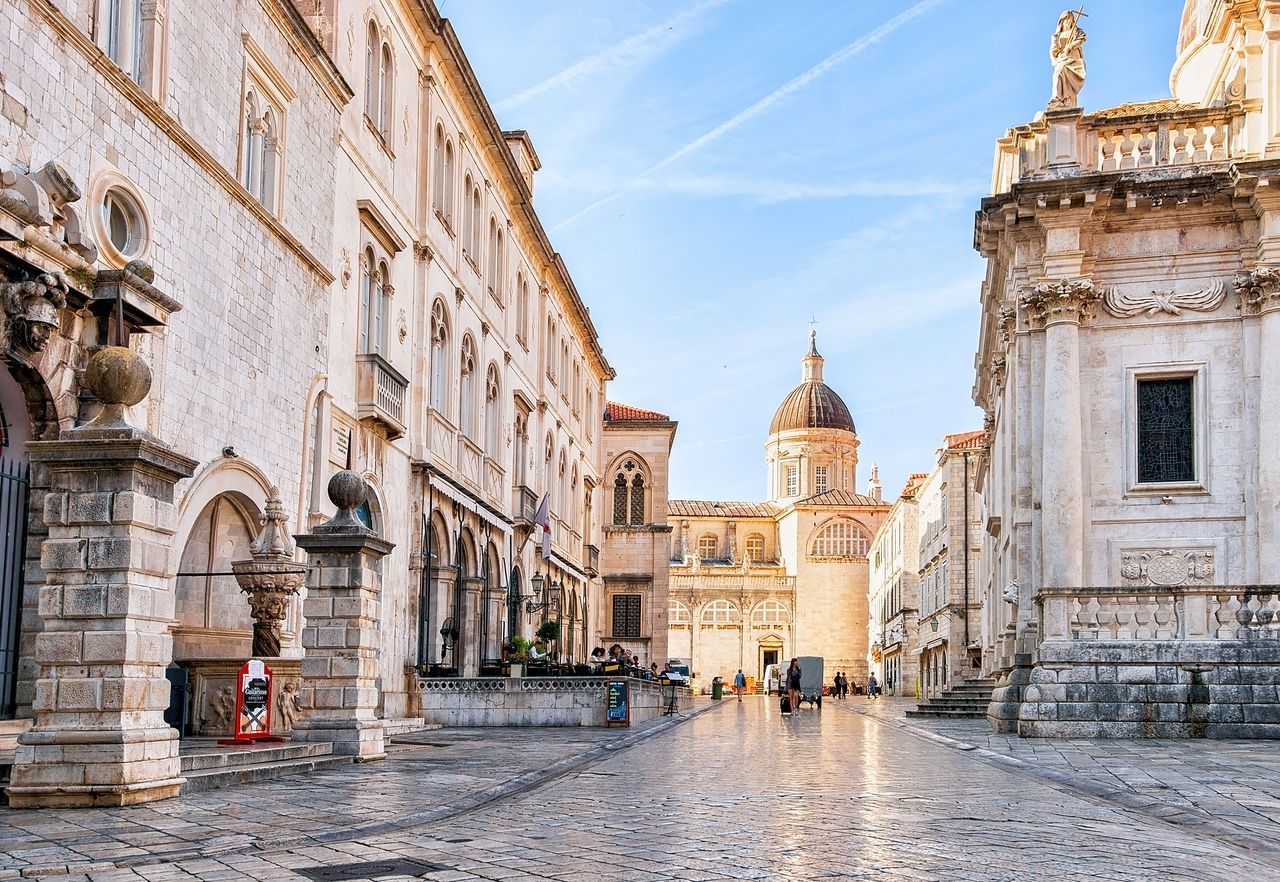 Een foto van een geplaveide straat in Dubrovnik, Kroatië, omgeven door historische gebouwen van lichte steen. Op de achtergrond is een kerk met een opvallende koepel te zien. De gevels van de gebouwen zijn versierd met decoratieve elementen en ramen. Enkele mensen wandelen langs de straat, terwijl de lucht licht bewolkt is.