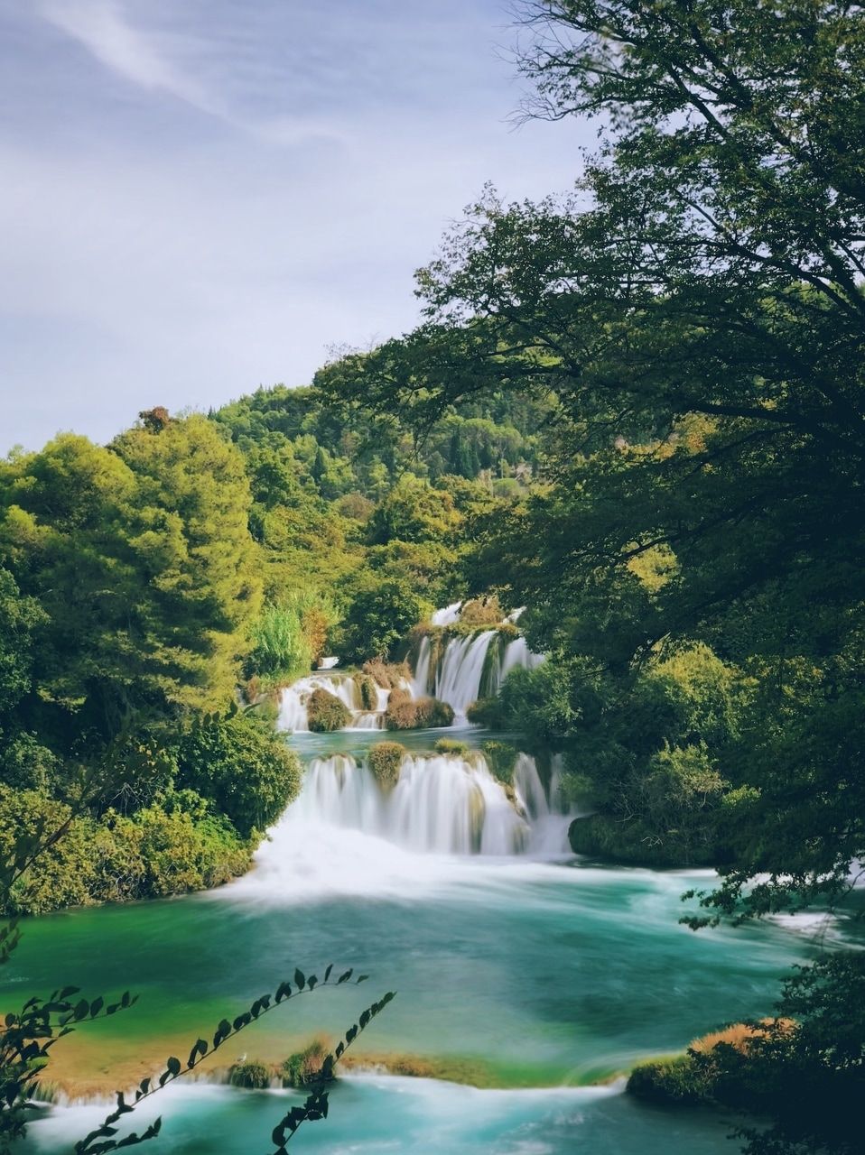 Cascadă în mijlocul unui peisaj forestier verde. O cascadă pitorească, înconjurată de copaci și plante luxuriante, verzi. Apa curge lin într-un bazin clar, de culoare turcoaz. Cerul este ușor înnorat și vegetația densă creează o atmosferă liniștită și conectată cu natura.