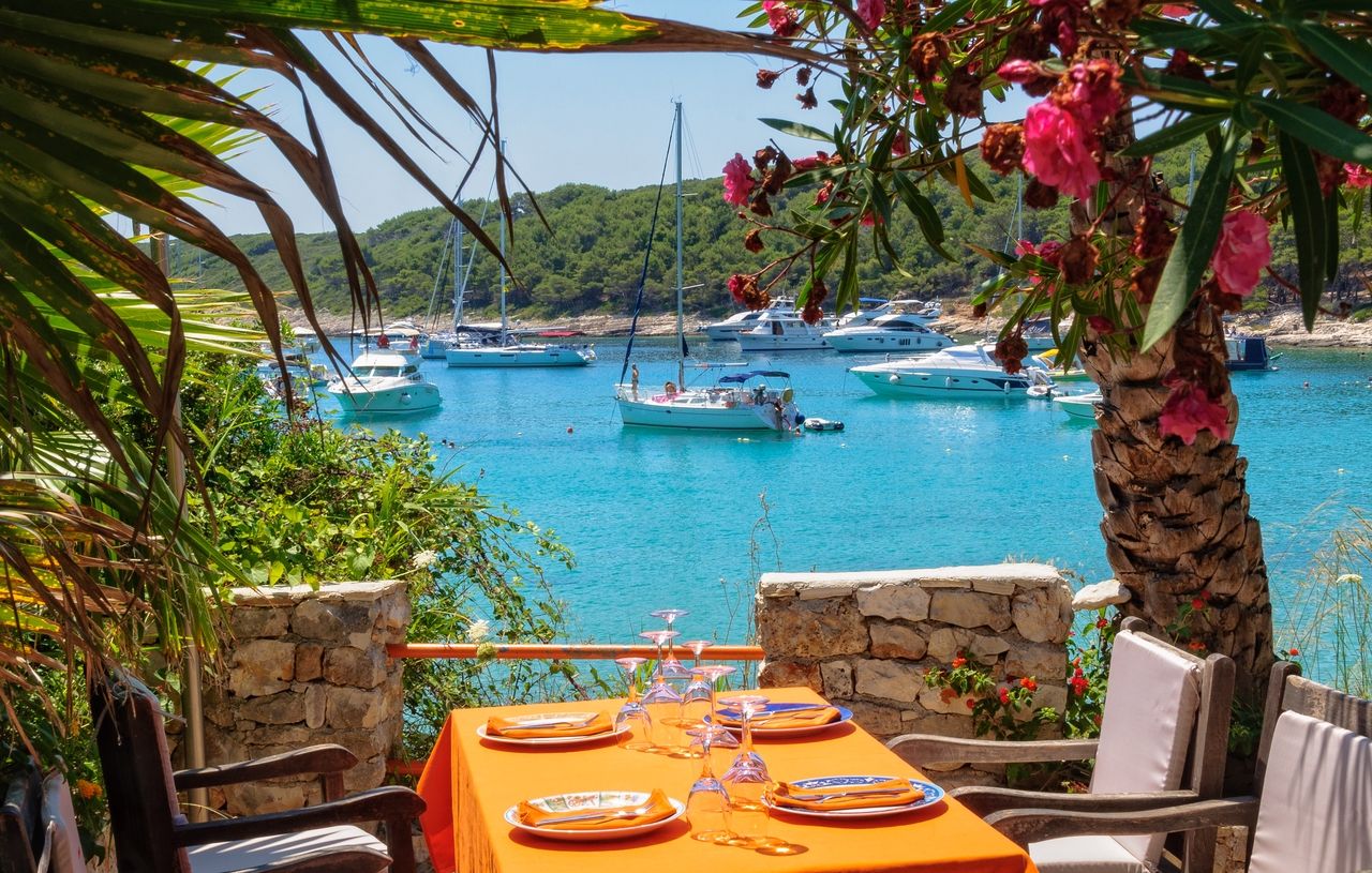 Ein idyllisches Essensarrangement im Freien mit Blick auf eine wunderschöne Bucht mit türkisfarbenem Wasser und zahlreichen Booten. Der Tisch ist mit einer orangefarbenen Tischdecke gedeckt und von üppigem Grün sowie blühenden Pflanzen umgeben.