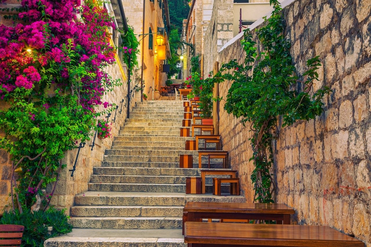 En brant stenstrappa i Hvar, Kroatien, kantad av färgglada blommor och frodig grönska. Längs trappan står små träbord och bänkar som skapar en mysig atmosfär. Byggnadernas väggar är av ljus sten och i bakgrunden syns lyktor som sprider ett varmt ljus. Scenen utstrålar en charmig, inbjudande stämning.