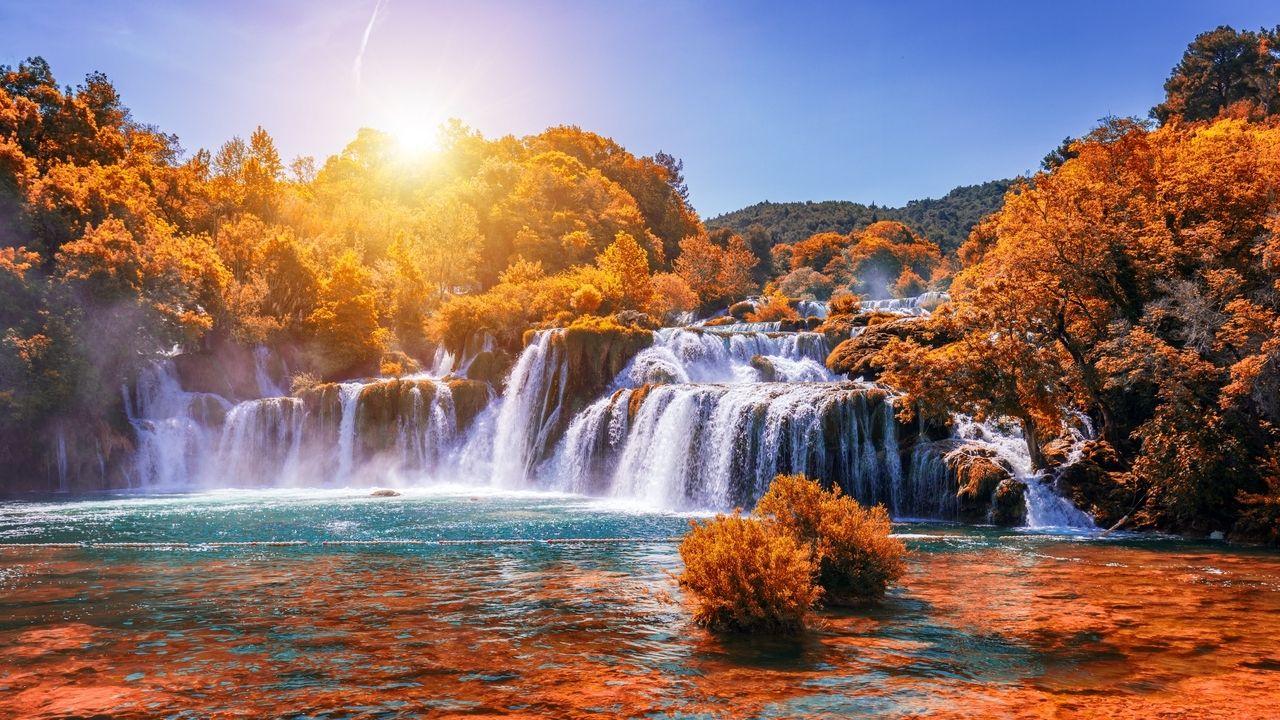 Le cascate nel Parco Nazionale di Krka in Croazia in autunno. L'acqua scende su diverse cascate in un fiume turchese, circondato da alberi dai colori autunnali brillanti. Il sole splende luminoso e immerge la scena in una luce calda e dorata, che rende il paesaggio colorato ancora più intenso.