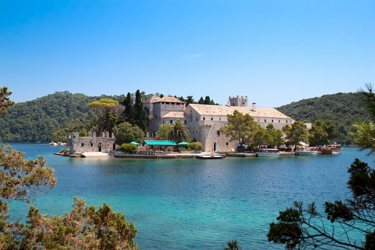 Eine malerische Ansicht des Benediktinerklosters auf der Insel Mljet, Kroatien. Das historische Kloster befindet sich auf einer kleinen Insel inmitten eines klaren, türkisfarbenen Sees. Umgeben von üppigem Grün und blauen Himmel, bietet das Kloster eine ruhige und idyllische Atmosphäre. Boote sind am Ufer festgemacht, und ein paar Menschen genießen die friedliche Umgebung.