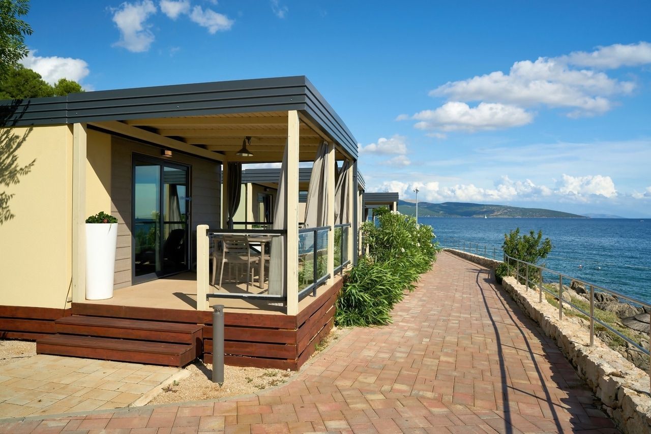 Bungalow moderno fronte mare. Un elegante bungalow con terrazza coperta e grandi finestre che offrono una vista sul mare blu. Un sentiero lastricato corre lungo la costa ed è fiancheggiato da piante verdi. Il cielo è sereno con poche nuvole, creando un'atmosfera rilassata e soleggiata.