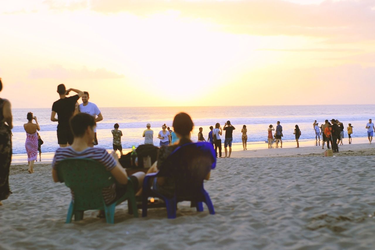 Menschen genießen einen wunderschönen Sonnenuntergang an einem Sandstrand. Einige sitzen auf Stühlen und entspannen, während andere am Ufer spazieren gehen und die friedliche Atmosphäre und die warme Abendsonne genießen.