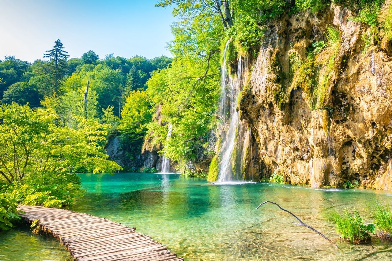 Der Nationalpark Plitvicer Seen in Kroatien mit einem hölzernen Wanderweg, der über kristallklares, türkisfarbenes Wasser führt, umgeben von üppiger grüner Vegetation. Im Hintergrund fließen mehrere Wasserfälle die Felswände hinunter in den See. Der Park strahlt natürliche Schönheit und eine friedliche, unberührte Atmosphäre aus.