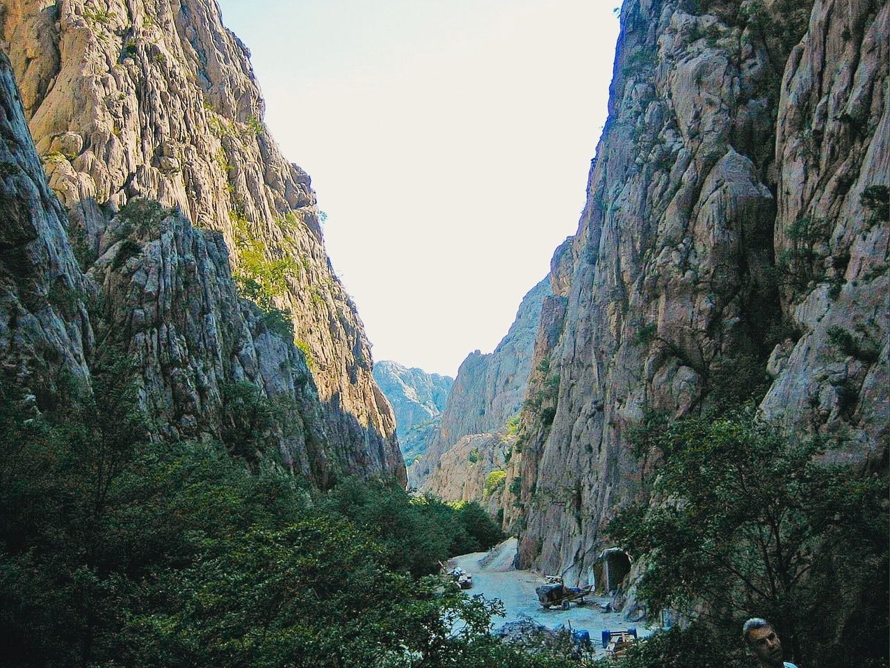 Canyon rocheux avec un sentier étroit. Un canyon impressionnant avec des parois rocheuses escarpées qui s'élèvent en hauteur. Au fond du canyon, un sentier étroit est entouré d'une végétation dense. À l'arrière-plan, on peut voir d'autres montagnes qui créent une formation paysagère dramatique et naturelle. Le ciel est clair, ce qui met en valeur les formations rocheuses majestueuses et la profondeur du canyon.