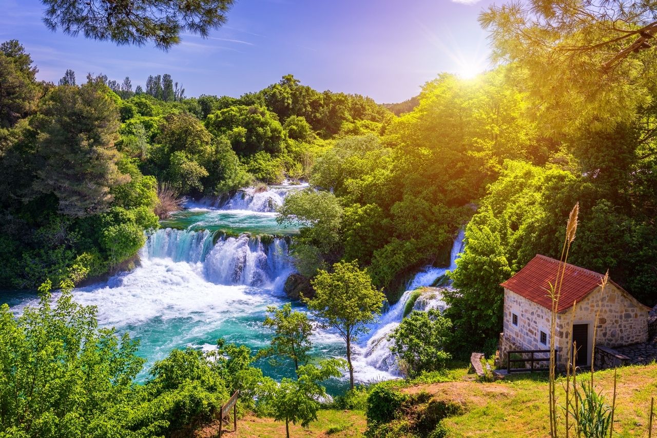 De watervallen van Roški Slap in Nationaal Park Krka in Kroatië. Het water stroomt in meerdere cascades door weelderig, groen loof en mondt uit in een turquoise rivier. Aan de rechterkant staat een klein stenen huis met een rood dak, omgeven door bomen. De zon schijnt fel en dompelt de scène in een warm, gouden licht, waardoor de natuurlijke schoonheid van de plek wordt benadrukt.