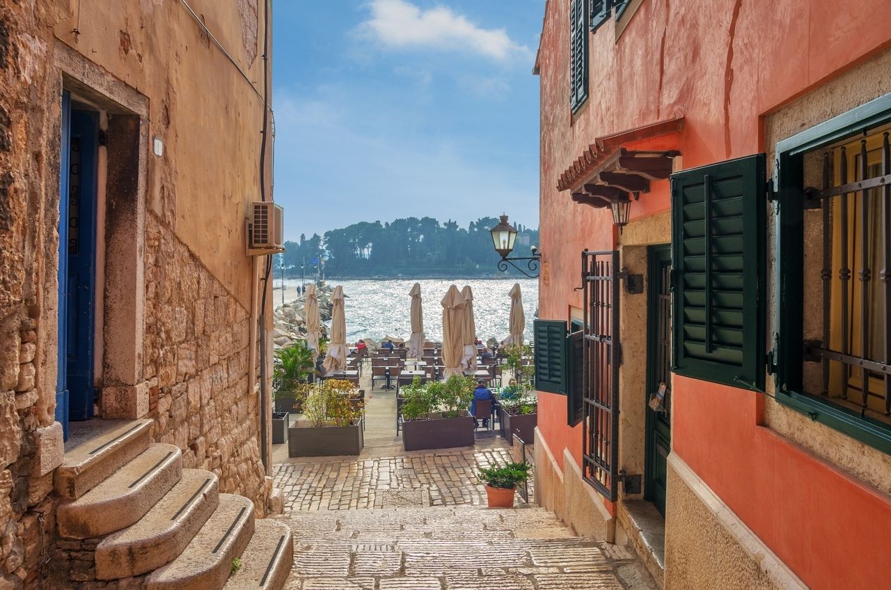 Un sentiero stretto e lastricato a Rovigno, Croazia, conduce a un'accogliente area esterna con tavoli e ombrelloni direttamente sul mare. A sinistra e a destra del sentiero ci sono edifici storici in pietra e intonaco colorato con persiane verdi. Sullo sfondo si estende l'acqua blu tranquilla, e all'orizzonte si vedono alberi e una piccola isola. La scena emana un'atmosfera mediterranea rilassata.