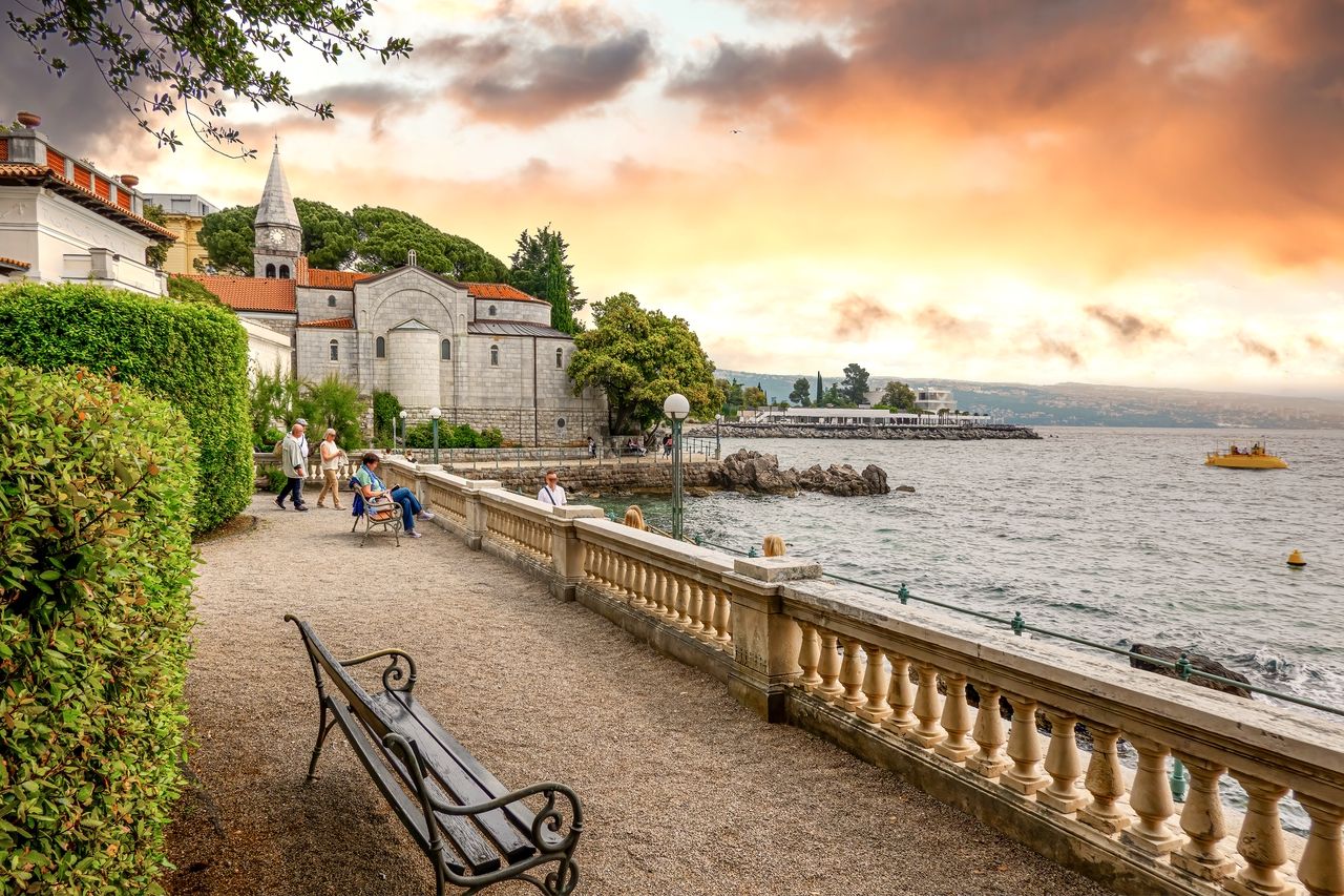 En pittoresk spaservei langs kysten i Opatija ved solnedgang. Folk nyter den avslappede atmosfæren, sitter på benker og spaserer langs stien. En historisk kirke med markant arkitektur og røde tak ligger på den ene siden, mens havet og kysten er synlig på den andre siden.