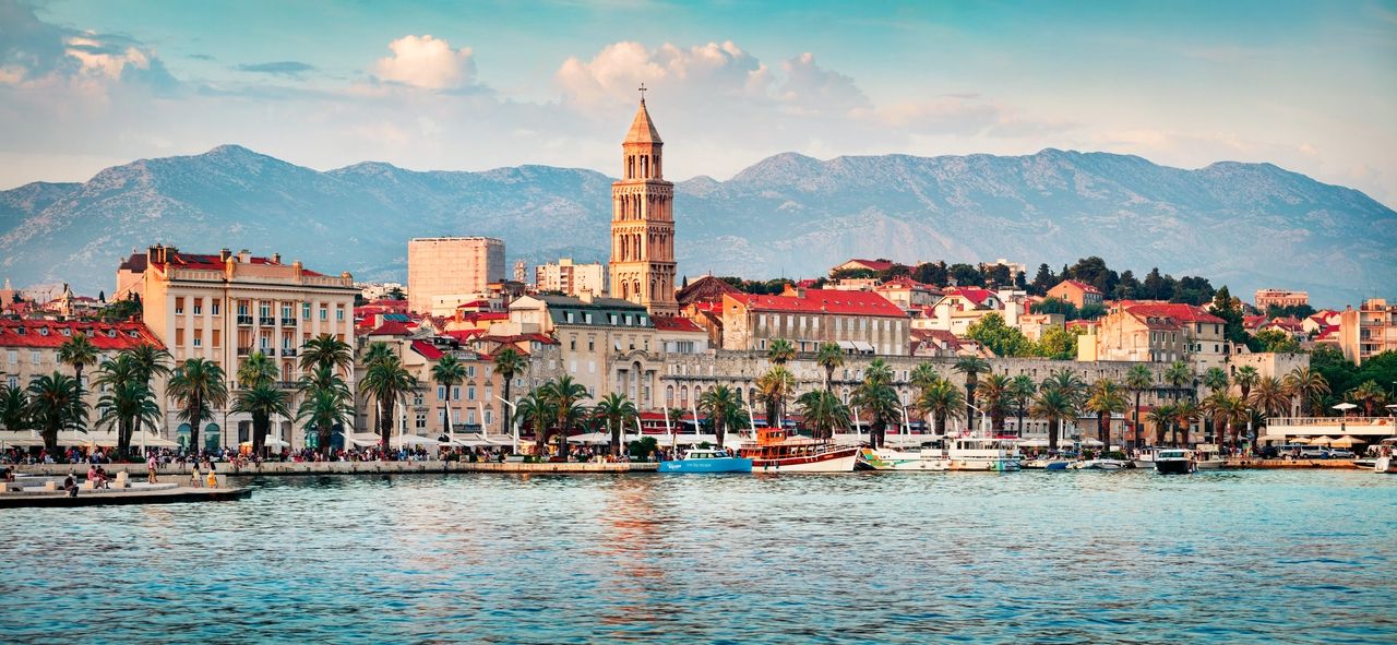 A photo of the Split coastline with a view of Diocletian's Palace and the waterfront promenade. Palm trees line the promenade, behind which historic buildings with red tile roofs rise. In the background, the distinctive mountains provide an impressive backdrop for the city. Boats and yachts are anchored in the clear blue waters of the harbor.