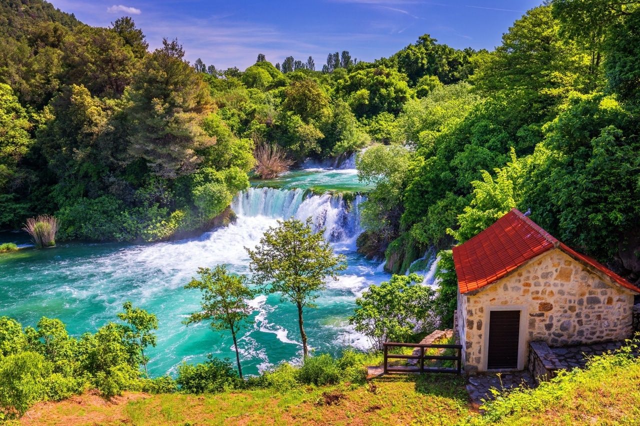 Idílico paisaje primaveral con cascada y pequeña casa de piedra. Una hermosa cascada rodeada de exuberante follaje verde. En primer plano hay una pequeña casa de piedra con techo de tejas rojas, incrustada en la naturaleza verde. El cielo azul claro completa la escena pacífica y transmite una sensación de tranquilidad y conexión con la naturaleza.