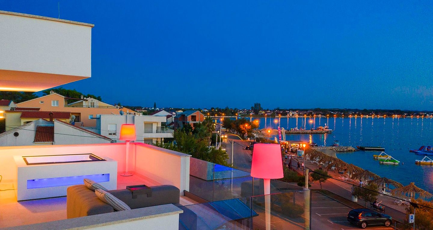 Ferienwohnung mit Meerblick in Kroatien bei Nacht. Moderne Terrasse mit stilvoller Beleuchtung, Blick auf die Stadt und das Meer mit beleuchteten Booten und Stränden. Ideal für einen entspannten Sommerurlaub.