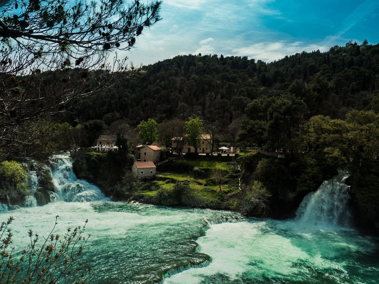 Village avec cascade au milieu d'une forêt. Un village idyllique avec des maisons en pierre traditionnelles est niché dans un paysage forestier verdoyant. Deux cascades impressionnantes traversent le village, entourées de végétation luxuriante et de grands arbres. Le ciel est partiellement nuageux, créant une atmosphère calme et naturelle.