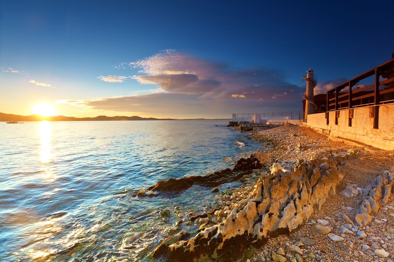 Zalazak sunca na obali Zadra, Hrvatska. Sunce zalazi iza planina i obasjava more zlatnom svjetlošću. Na desnom rubu slike nalazi se svjetionik sa spiralnim stubištem, smješten na stjenovitoj obali. Bistra voda svjetluca u različitim nijansama plave boje, a scena odiše mirnom i spokojnom atmosferom.