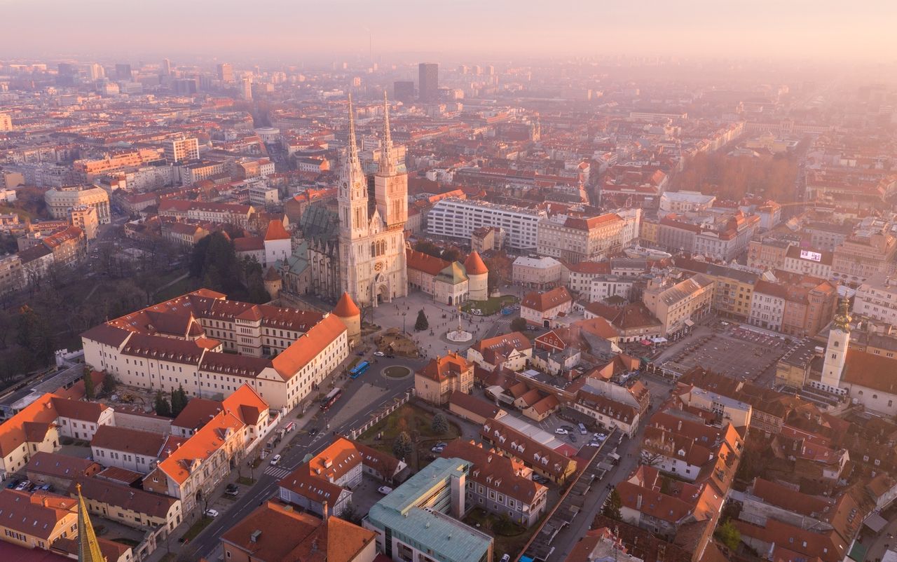 Luftaufnahme von Zagreb bei Sonnenuntergang mit Blick auf die Kathedrale von Zagreb. Die Kathedrale steht im Zentrum des Bildes und ist von historischen Gebäuden mit roten Dächern umgeben. Die Stadt erstreckt sich in die Ferne und ist in ein warmes, goldenes Licht getaucht, das die Gebäude und Straßen sanft beleuchtet. Der Himmel ist klar, und die Stadt wirkt ruhig und friedlich.