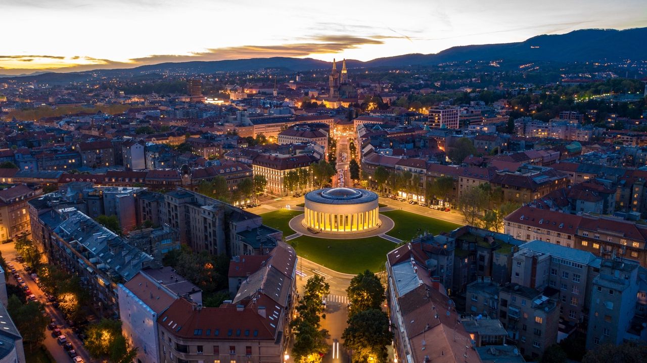 Vista aérea de Zagreb al atardecer. En el centro de la imagen se encuentra el edificio iluminado del Teatro Nacional de Croacia en una gran plaza, rodeado de árboles y calles iluminadas. La ciudad se extiende hacia el fondo, con la silueta distintiva de la Catedral de Zagreb en el horizonte. Las montañas forman un pintoresco telón de fondo a la luz del atardecer.