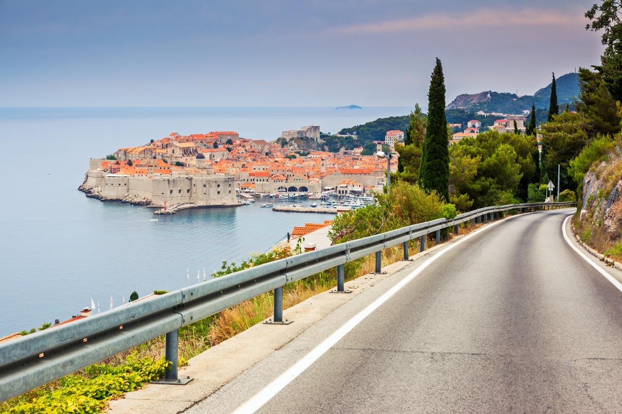 Panoramautsikt over den historiske gamlebyen i Dubrovnik i Kroatia, sett fra en svingete vei som fører ned til kysten. De antikke bymurene og røde takene står i kontrast til det klare, blå havet og den omkringliggende frodige vegetasjonen.