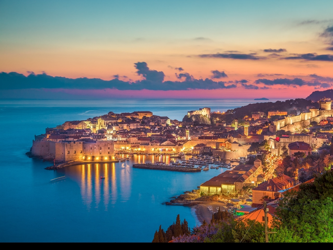 Opplyst gamleby i Dubrovnik i Kroatia ved solnedgang. De historiske bymurene og bygningene speiler seg i det rolige vannet i Adriaterhavet og skaper en romantisk og pittoresk atmosfære.