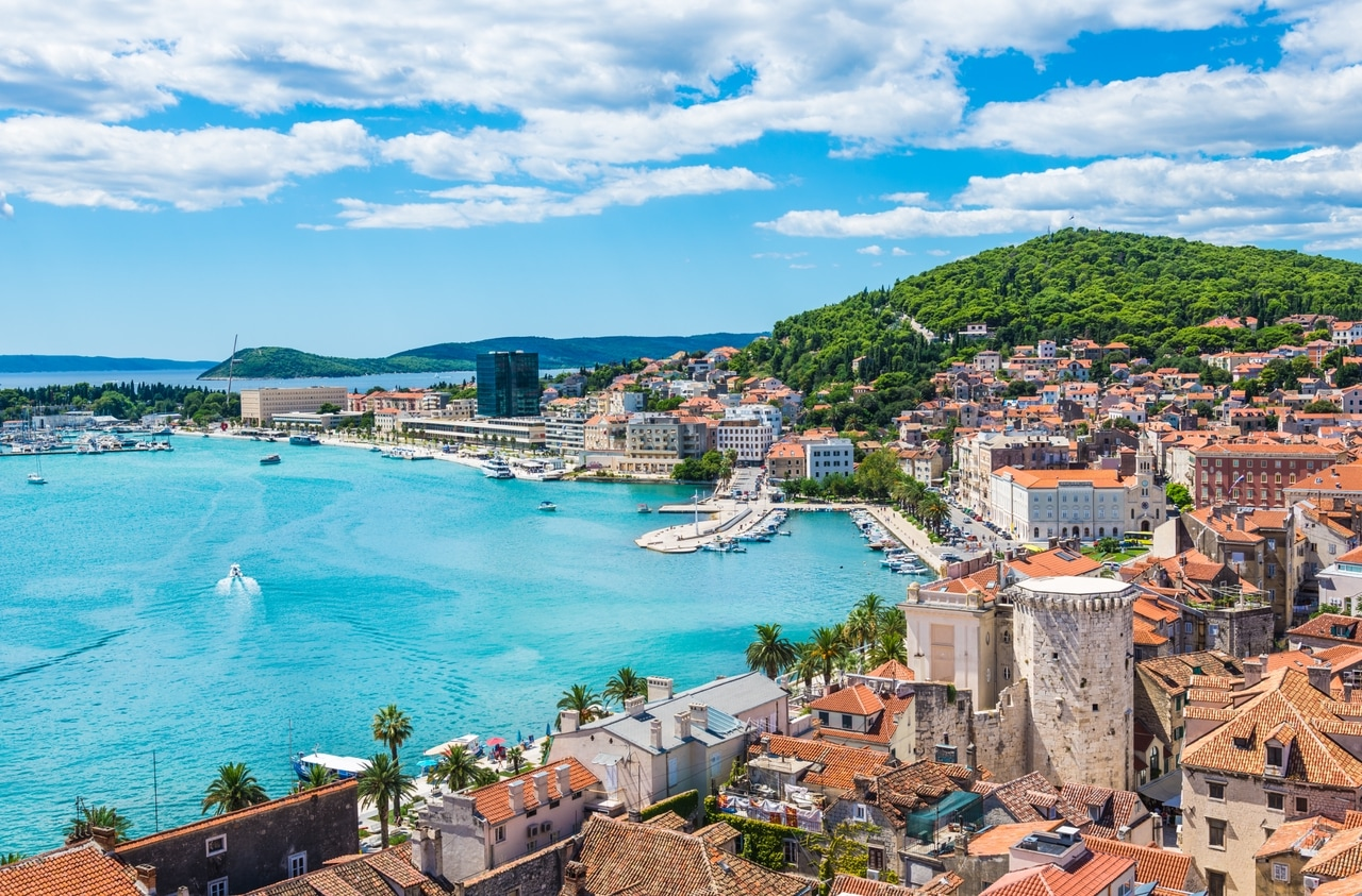 Panorámakép Split tengerparti városáról Horvátországban, történelmi belvárosával, festői kikötőjével és a környező zöld dombokkal. A tiszta, kék Adria és a bájos építészet Splitet népszerű úti céllá teszik.