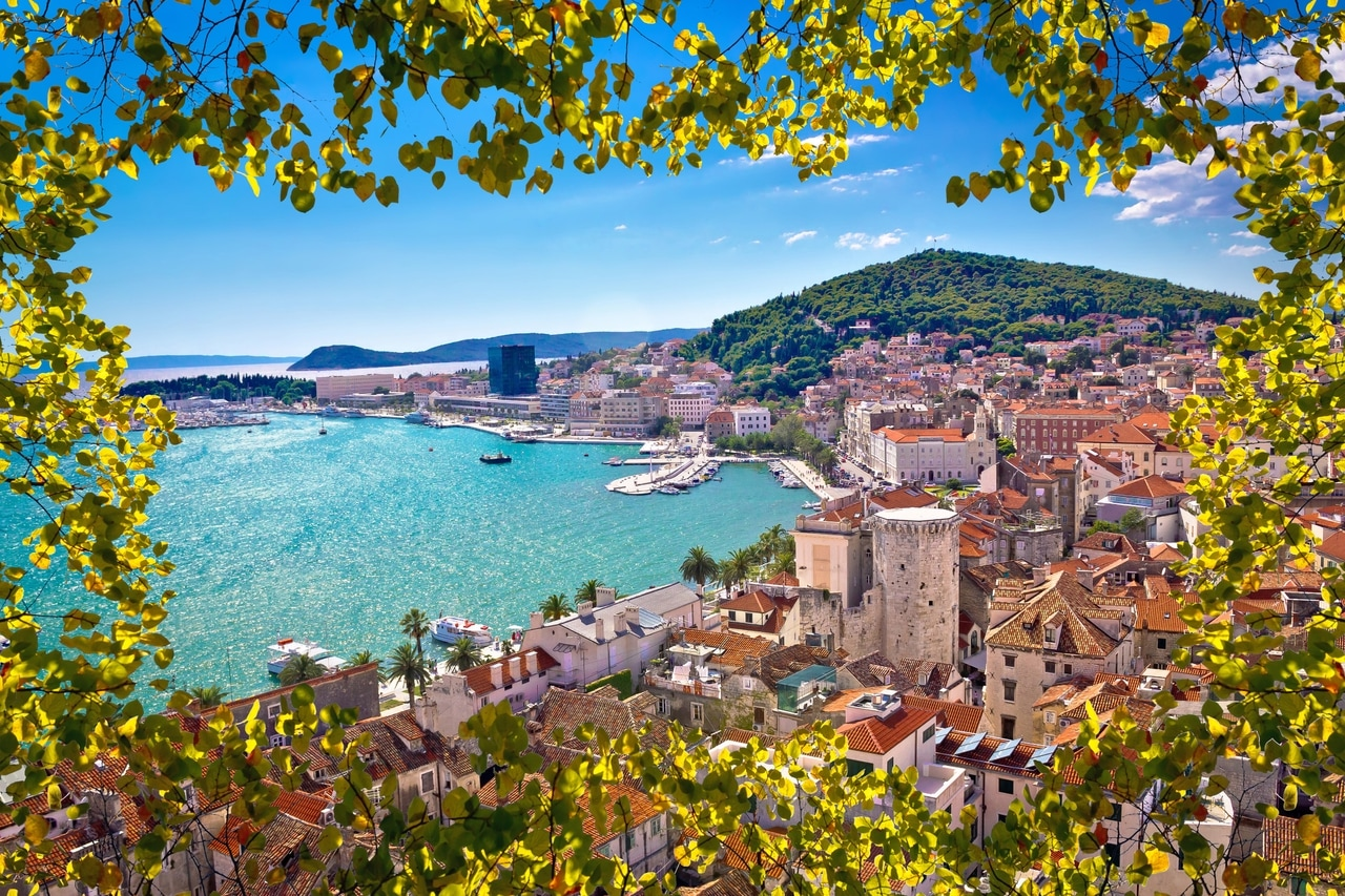 Panoramski pogled na obalni grad Split u Hrvatskoj, uokviren zelenim lišćem. Povijesne zgrade, slikovita luka i okolna brda pružaju zadivljujući krajolik pod vedrim, plavim nebom.