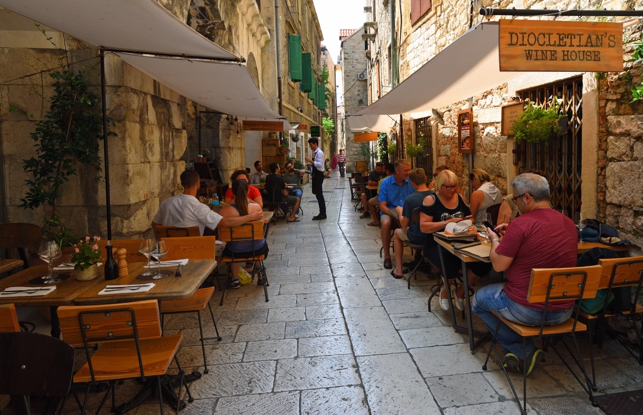 Scenă animată de stradă în orașul vechi din Split, Croația, cu oameni care stau într-un restaurant confortabil în aer liber. Străzile înguste, pavate și clădirile istorice creează o atmosferă fermecătoare și primitoare.