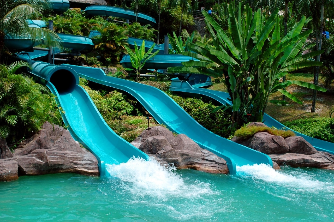 Aufregende Wasserrutschen in einem tropischen Wasserpark, umgeben von üppiger Vegetation und Palmen. Die hellblauen Rutschen enden in einem erfrischenden Pool, der ein perfektes Ziel für Spaß und Abenteuer bietet.