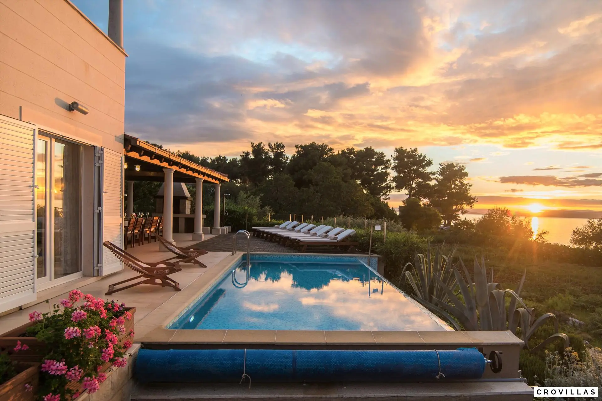 Luxuriöse Villa auf der Insel Brac mit Pool und Meerblick bei Sonnenuntergang, umgeben von grüner Vegetation und blühenden Pflanzen. Die Terrasse bietet Liegestühle und einen Essbereich im Freien, ideal für entspannte Abende.